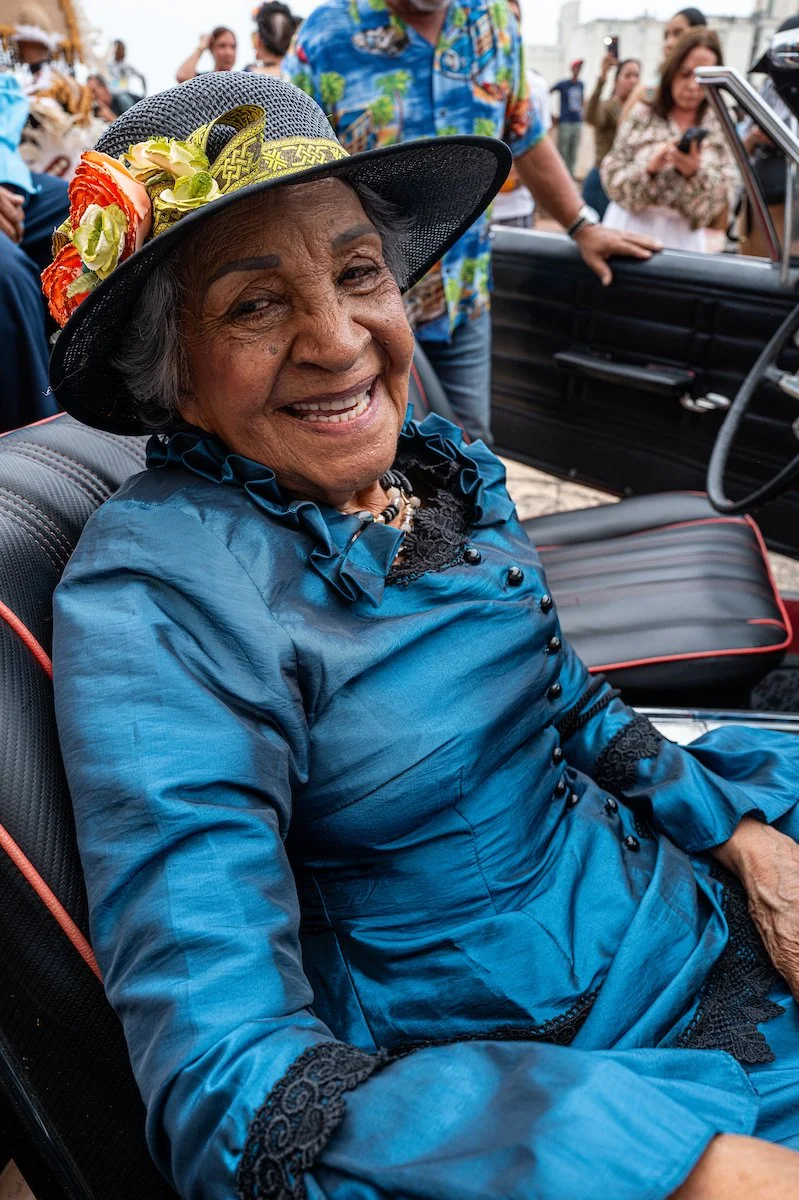 Smiling elderly woman in a blue dress and floral hat sitting in a vintage car at an outdoor event.