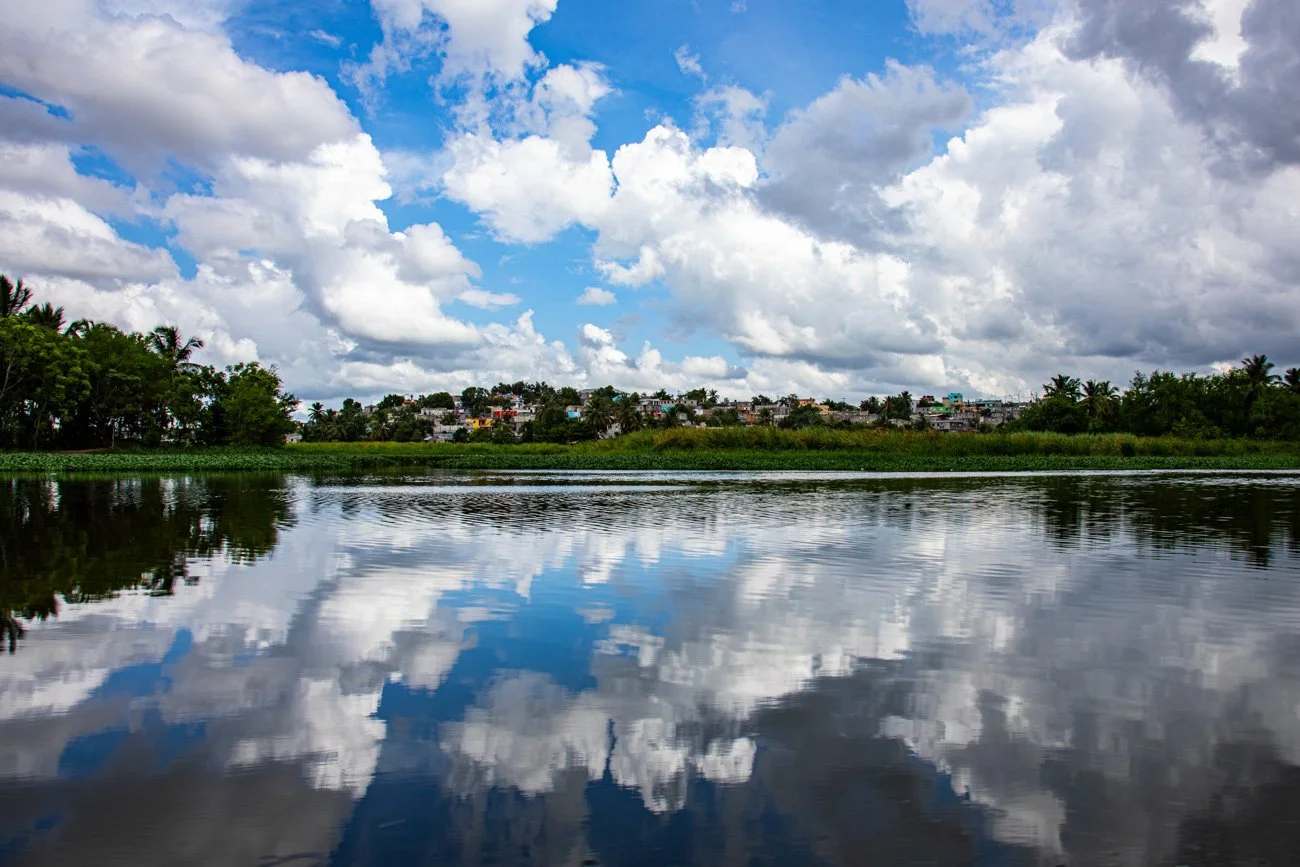 Cloudy sky reflecting on a calm body of water with greenery and buildings in the background.
