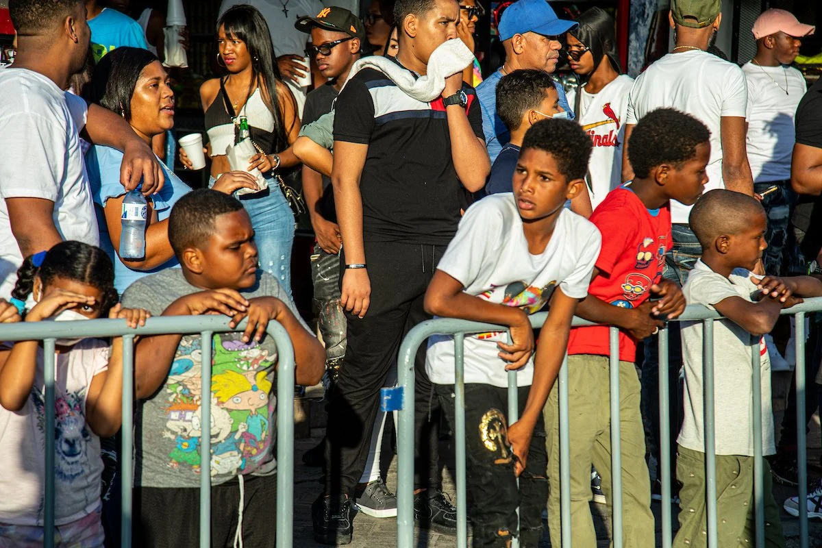 Group of children and adults standing behind a metal barrier at an outdoor event, some looking bored or distracted.