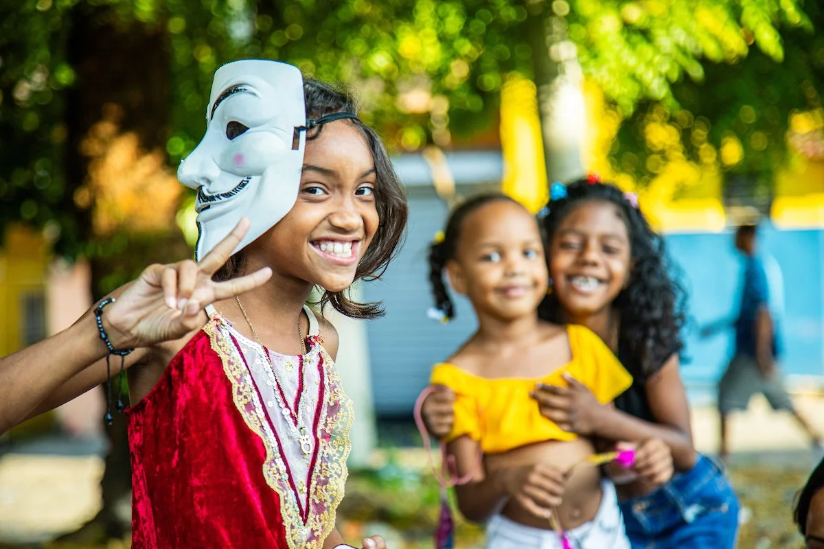 Child girl smiling and holding a mask over her eye at an outdoor gathering with other children.