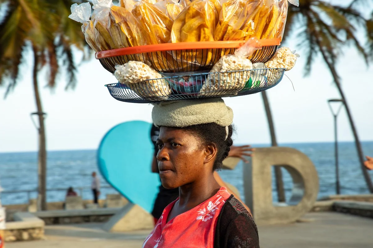 A woman balancing a large basket with food items on her head, standing near the ocean with palm trees in the background.
