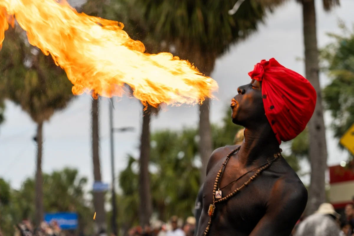 A man with dark skin and a red turban on his head breathes fire during a street performance, with trees and a crowd in the background.