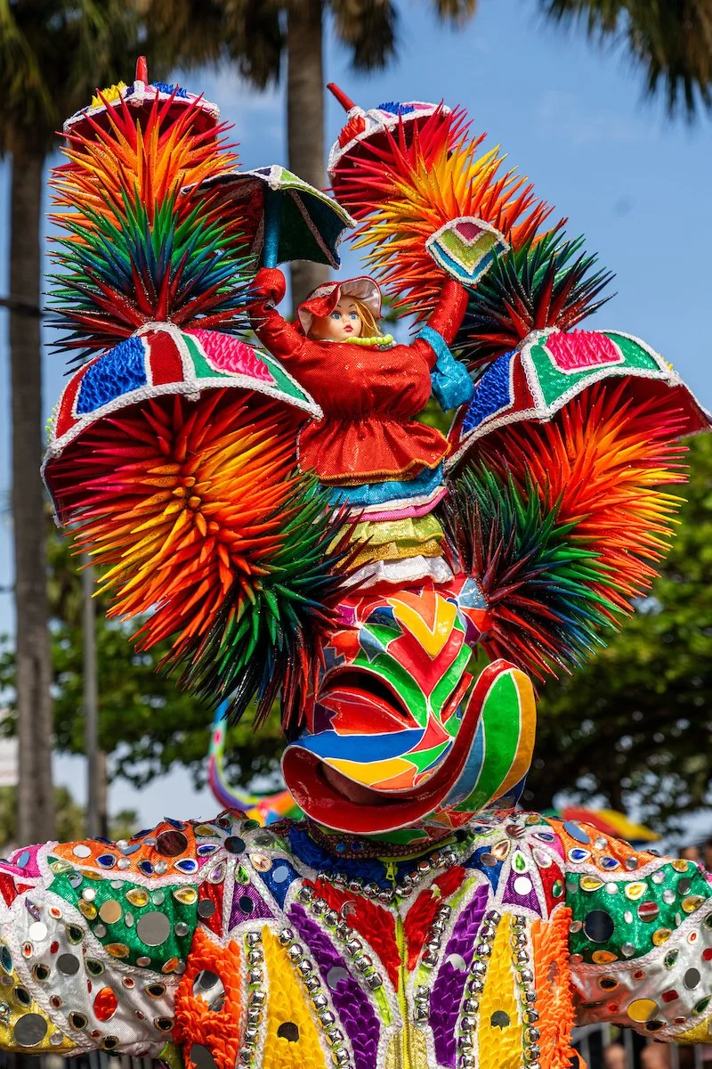 Colorful decorated figure with a smile, spiky headdress, and vibrant clothing, outdoors with trees and blue sky in the background.