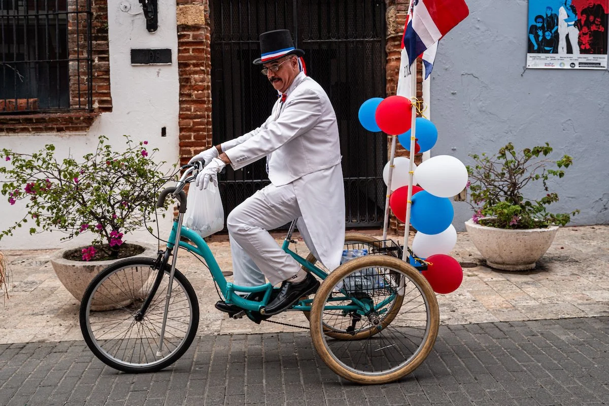 A man dressed as Uncle Sam riding a three-wheeled bicycle decorated with red, white, and blue balloons and flags, in a street with potted plants and posters on the wall.