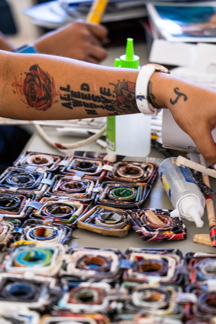 Close-up of a person's arm with tattoo art, jewelry, and a white wristband, reaching over a table with various colorful camera-shaped objects, modeling glue, and a green marker, in a creative workspace.