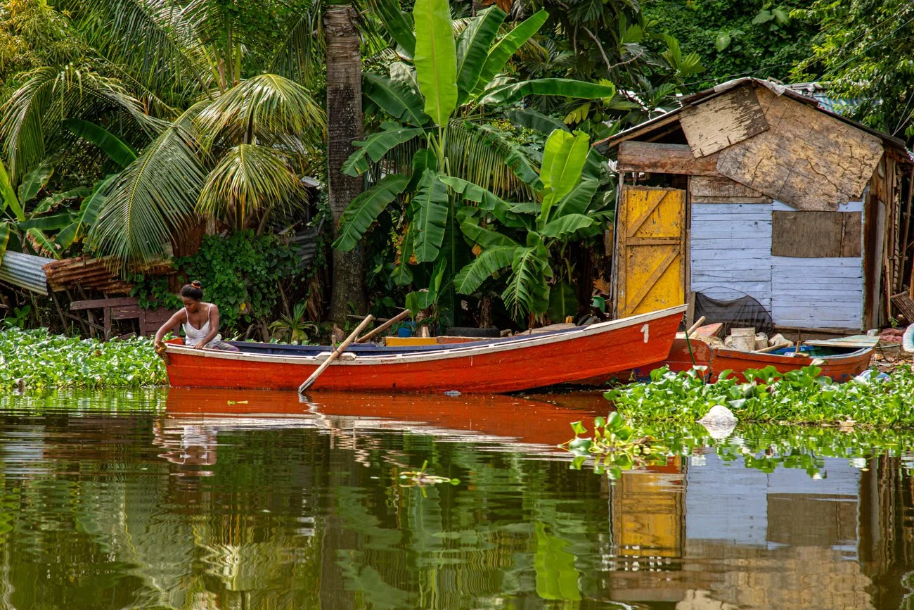 A woman in a white tank top is sitting in a small red boat on a river surrounded by dense green tropical plants and trees, with a makeshift wooden house nearby.
