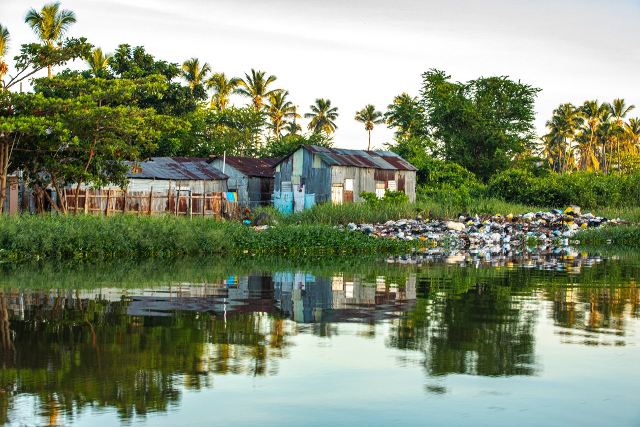 A rural scene with a small, old, rusty metal house surrounded by greenery and palm trees, next to a body of water with garbage floating on the surface.