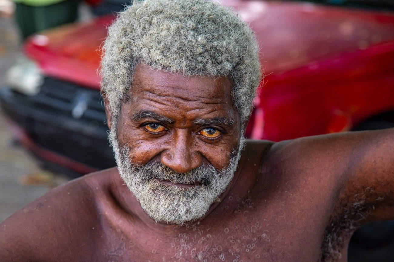 Close-up of an elderly man with gray hair and beard, looking directly at the camera with a serious expression. He is shirtless, and a red vehicle is blurred in the background.