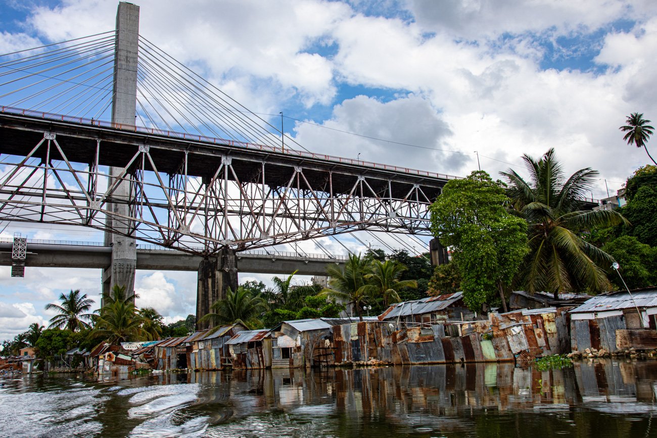 A large bridge above a waterfront shantytown with makeshift metal shacks and lush green trees, under a partly cloudy sky.