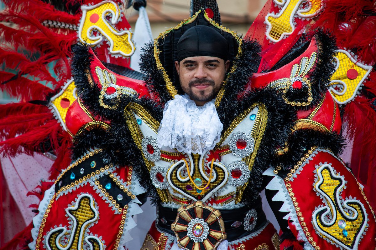 A man wearing an elaborate costume with red, black, white, and gold details, adorned with feathers, rhinestones, and embroidery, possibly for a cultural or festive event.