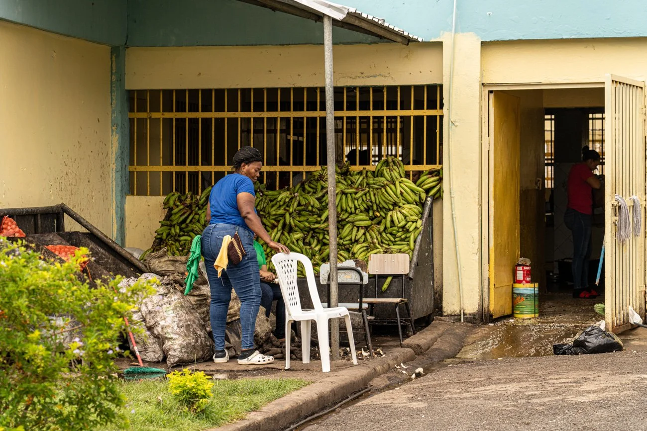 A woman in a blue shirt and jeans standing next to a chair, with a large pile of green bananas in the background outside a building. Inside, another woman appears to be working near a yellow door.