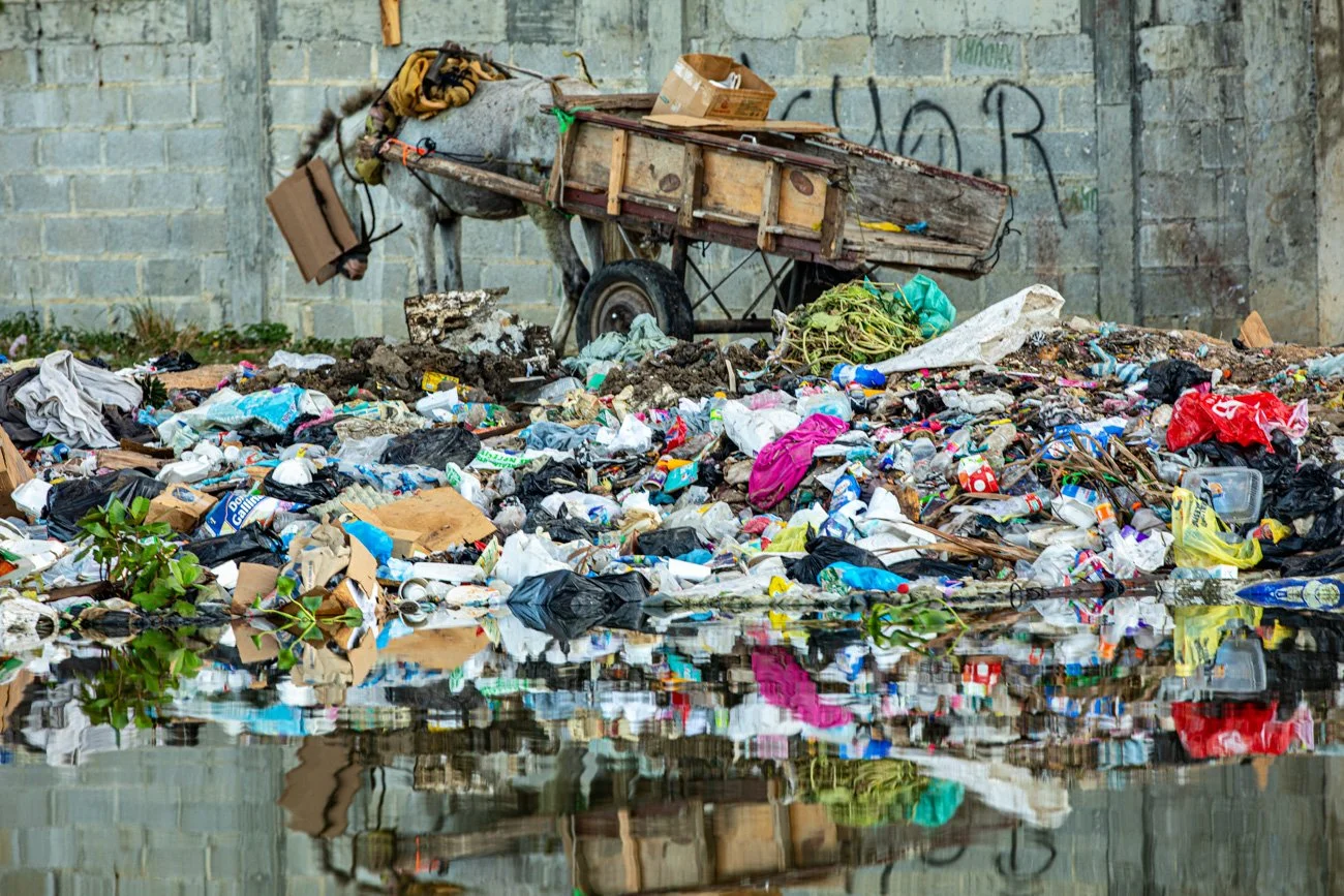 Overflow of trash and garbage on the ground near a stone wall, with a rusty wheelbarrow on the back containing bags and items, and a reflection of the trash in nearby water.