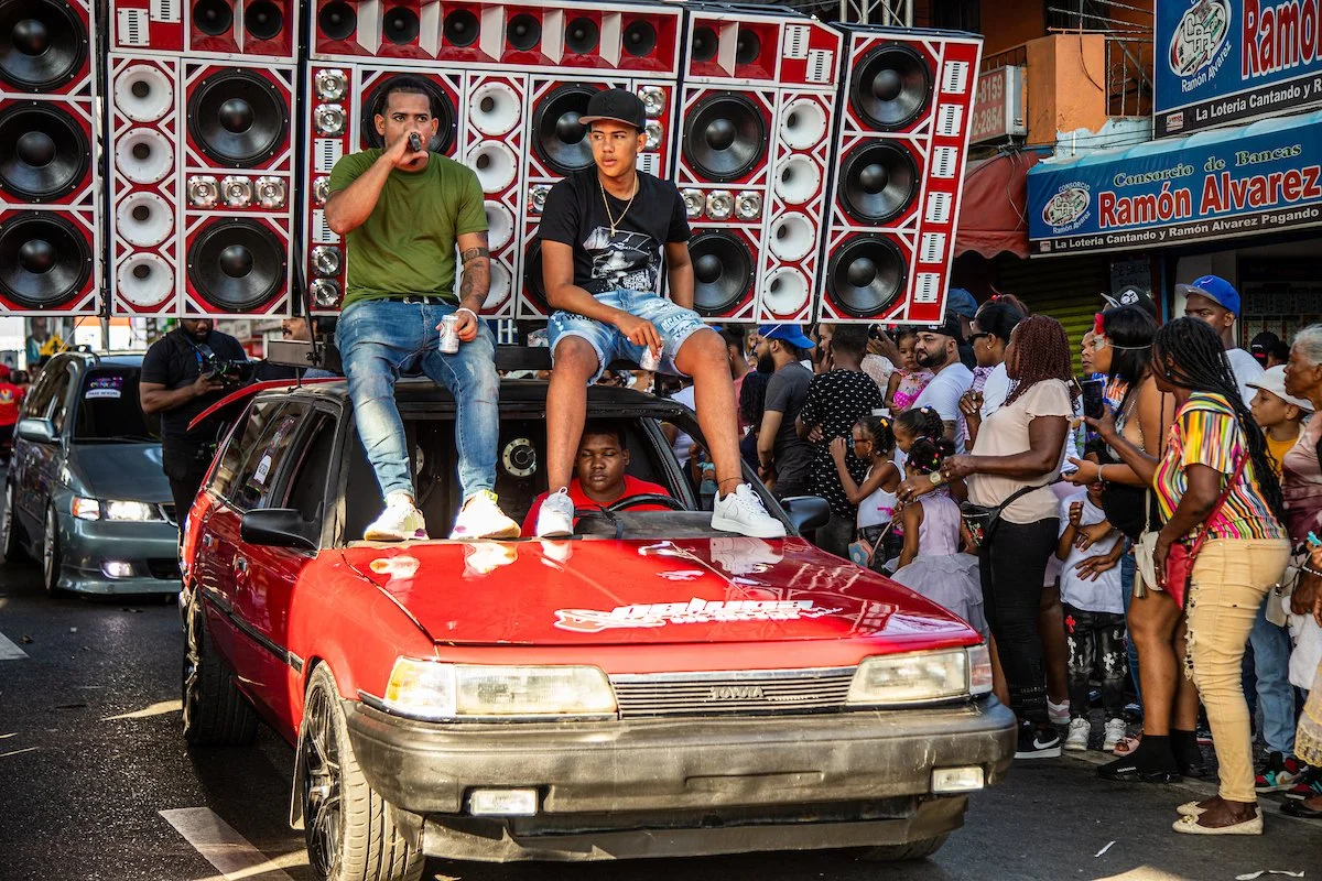 Two young men are sitting on the hood of a red car with large speaker systems attached to a truck above them. They are surrounded by a crowd of people on the street, some taking photos and watching the event.