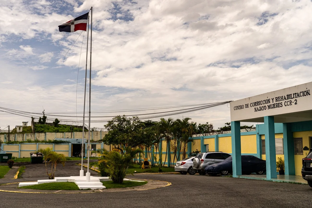 A parking lot outside a building with a sign that reads 'Centro de Corrección y Rehabilitación Najayo Mujeres CCR-2'. Four cars are parked near the building, with a flagpole displaying the Dominican Republic flag, green palm trees and bushes, and a p