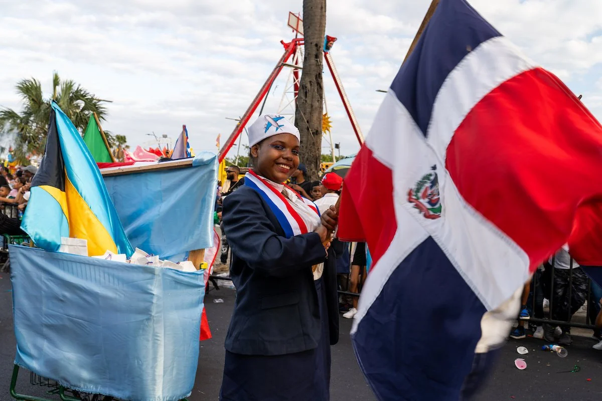 A young girl dressed in a dark blazer and honors sash, smiling and holding a small flag, at a parade or festival with flags and a Ferris wheel in the background.