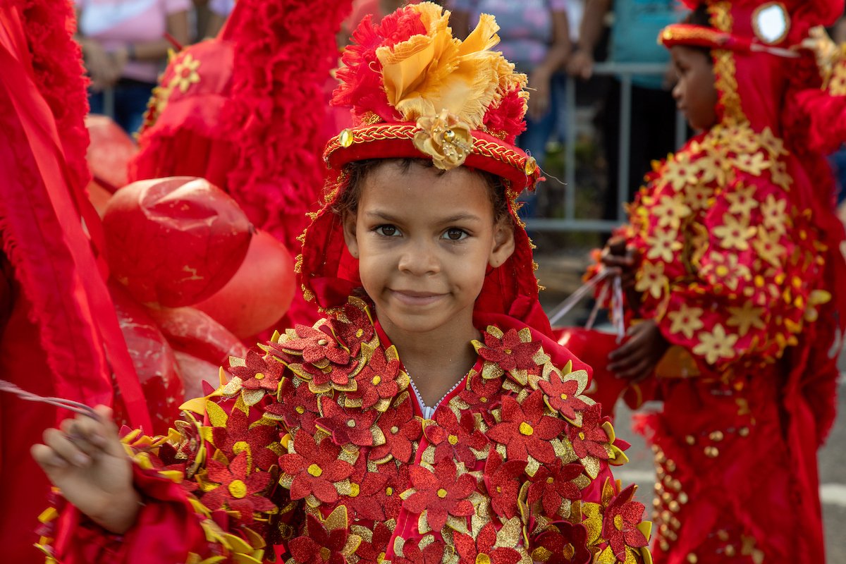 A young girl dressed in a vibrant red and gold costume with flower details, attending a festival or parade.