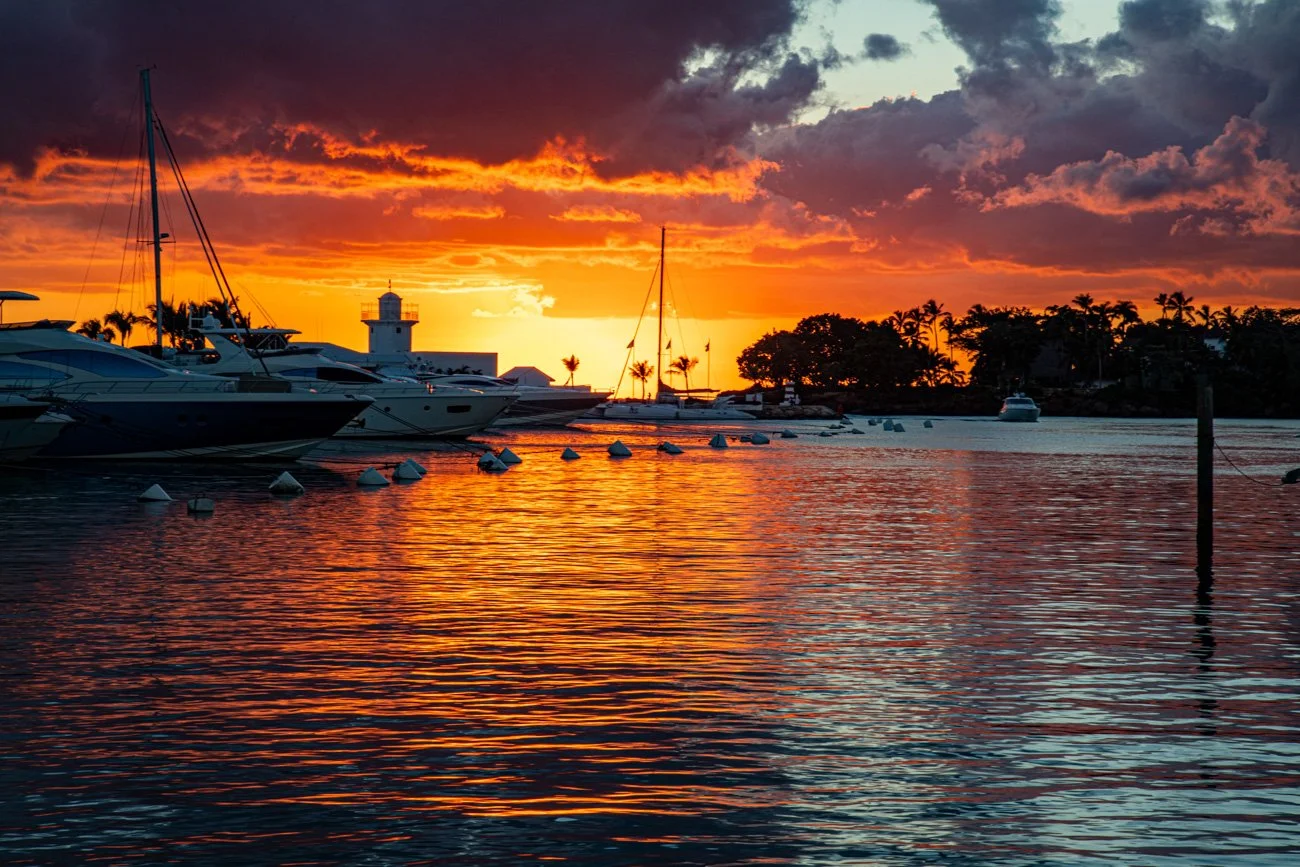 Boats docked at a marina during sunset, with orange and purple clouds reflected on the water and silhouettes of trees and a lighthouse in the background.