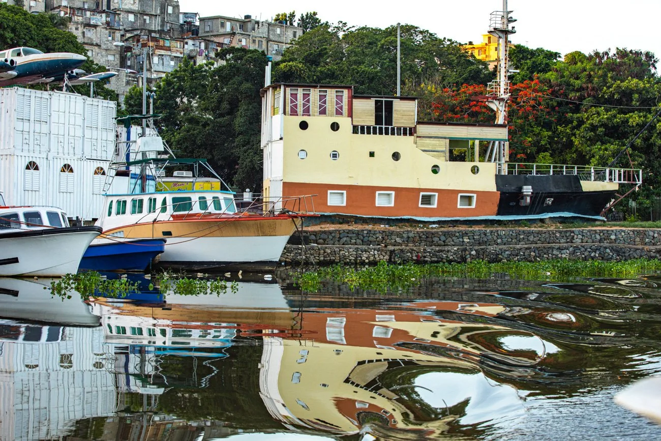 Boats docked along the canal with a houseboat designed like a ship, colorful buildings, and greenery in the background. Reflection of boats and buildings in the water.
