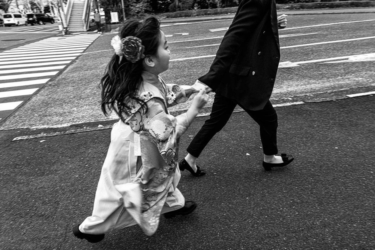 A young girl wearing traditional Japanese clothing holds hands with an adult crossing a street.