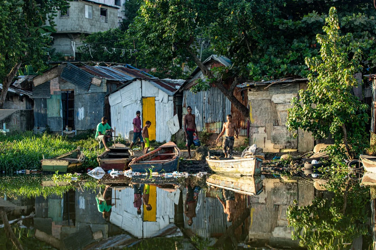 A group of young men by the water near a makeshift shoreline in a neighborhood with dilapidated houses, some of which are made of rusted metal sheets, surrounded by green trees and vegetation.