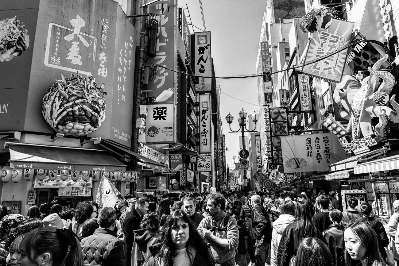 Crowded street scene in Japan with many people walking, various signs and advertisements in Japanese, and a large decorative octopus on a sign, with overhead banners and streetlights.