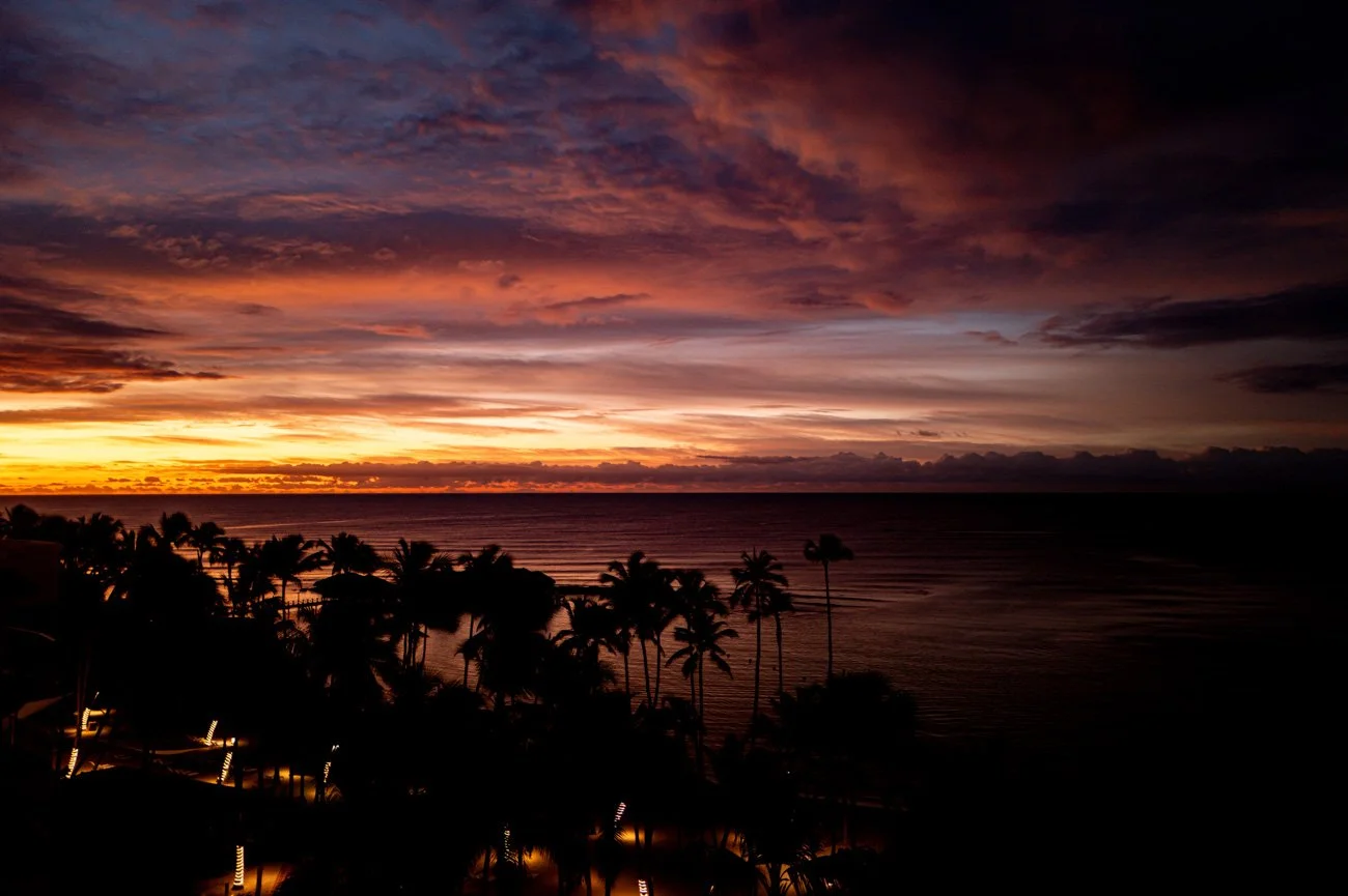 Sunset over the ocean with colorful clouds in the sky, silhouetted palm trees and illuminated outdoor area in the foreground.