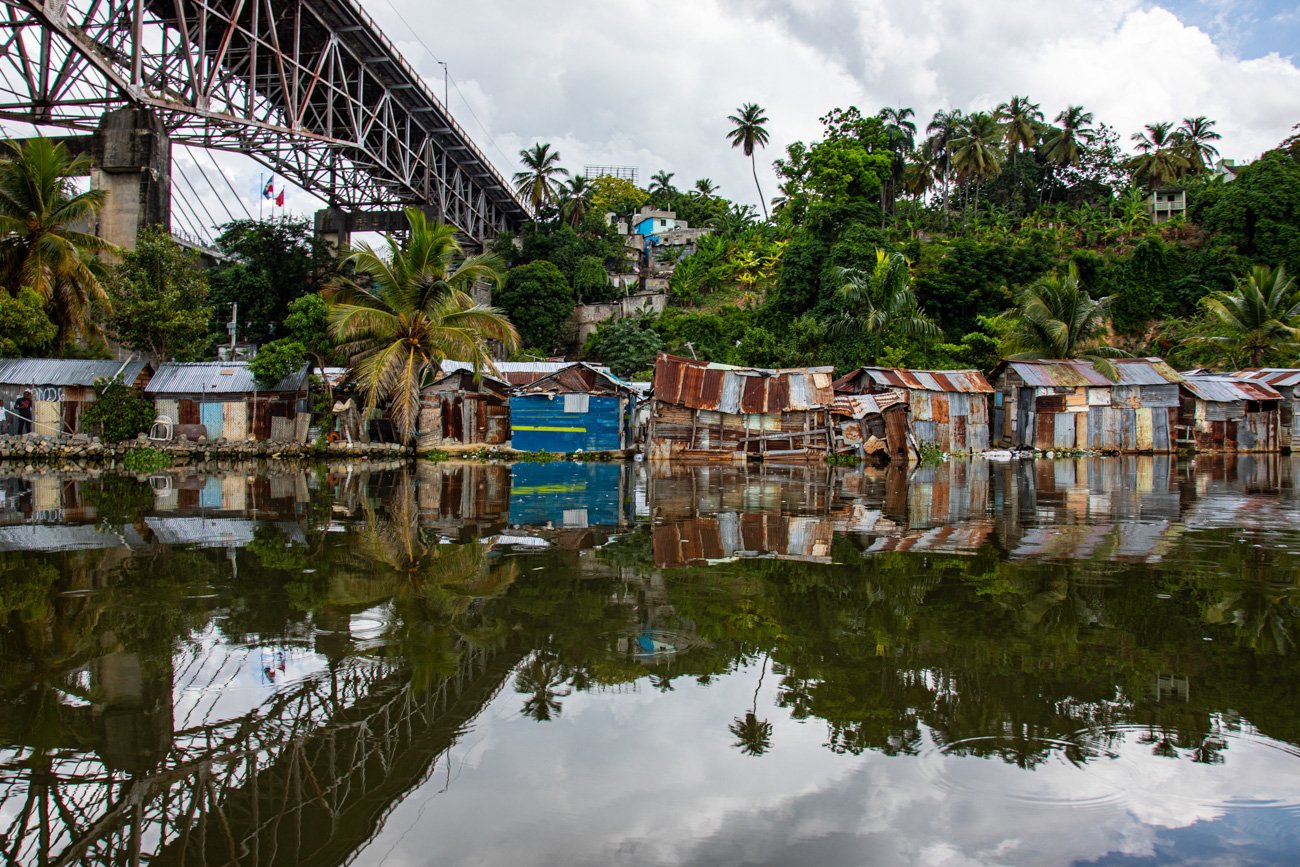 Dilapidated houses with rusted metal roofs along a waterway, reflections of houses and trees in the water, lush greenery, palm trees, and a bridge overhead under a cloudy sky.