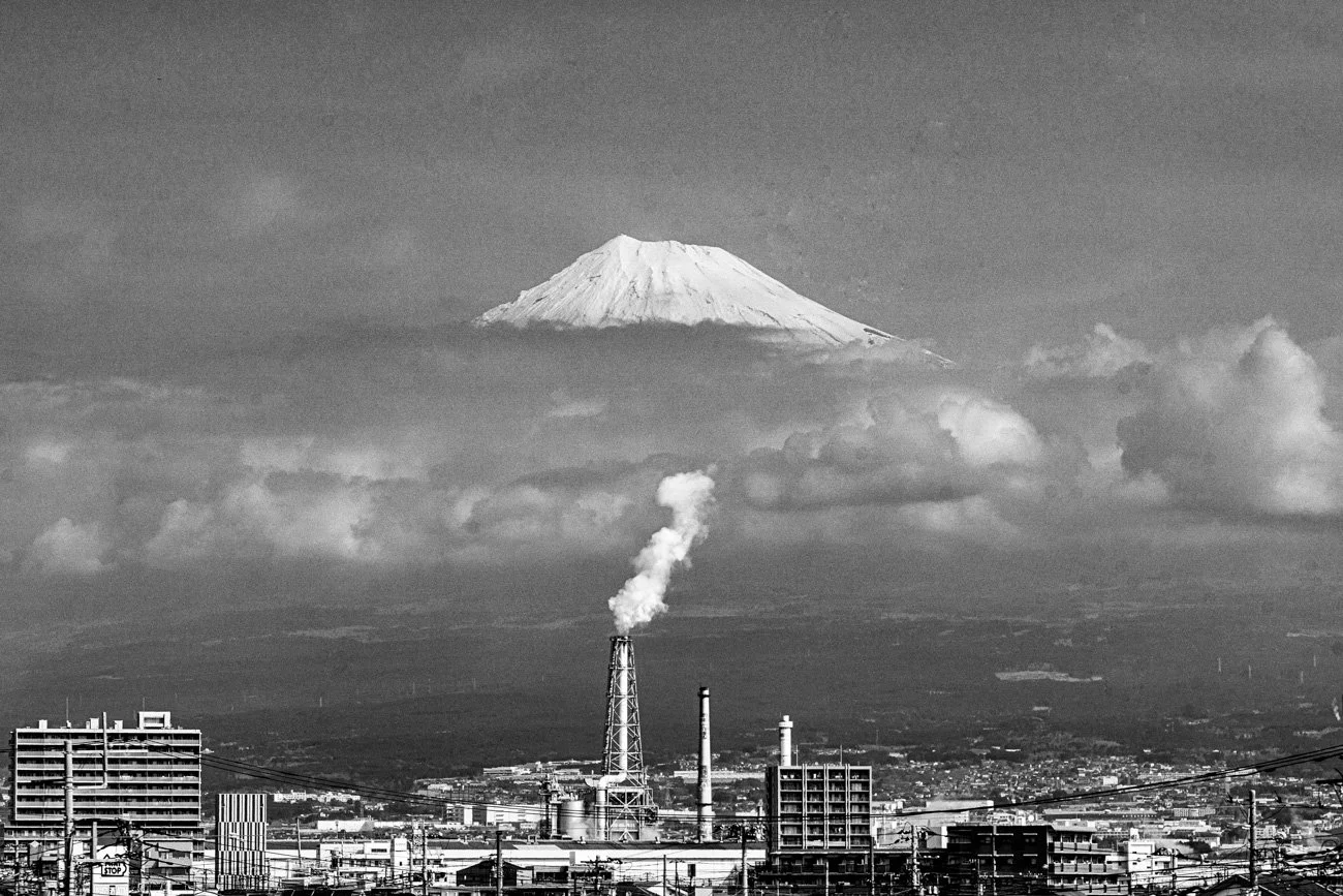 Black and white photo of a cityscape with industrial buildings and smokestacks in the foreground, clouds above, Mount Fuji in the background with a snow-capped peak.
