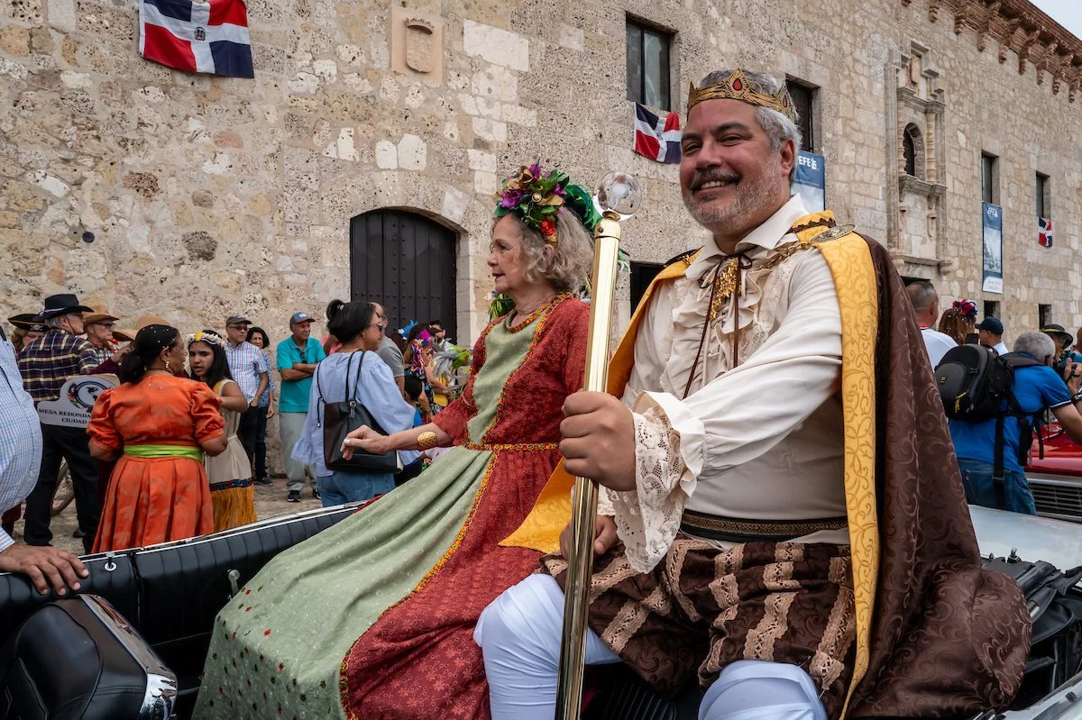 A man and a woman dressed in historical costumes riding in a decorated carriage during a parade. The man is wearing a crown and holding a scepter, and the woman is wearing a floral crown. There are people dressed in traditional clothing and observing