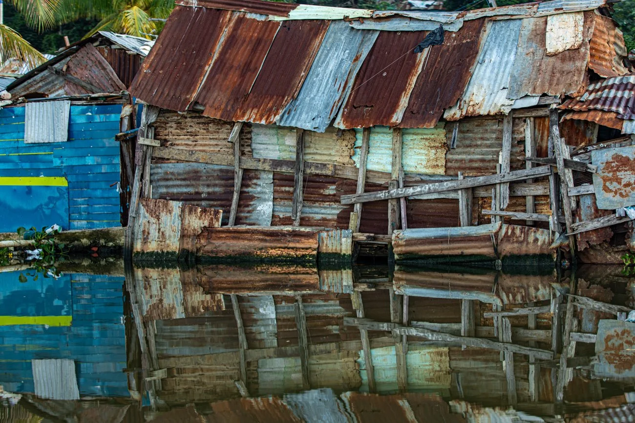 A dilapidated wooden and metal-shingled house. It is reflected in a body of water in front of it.