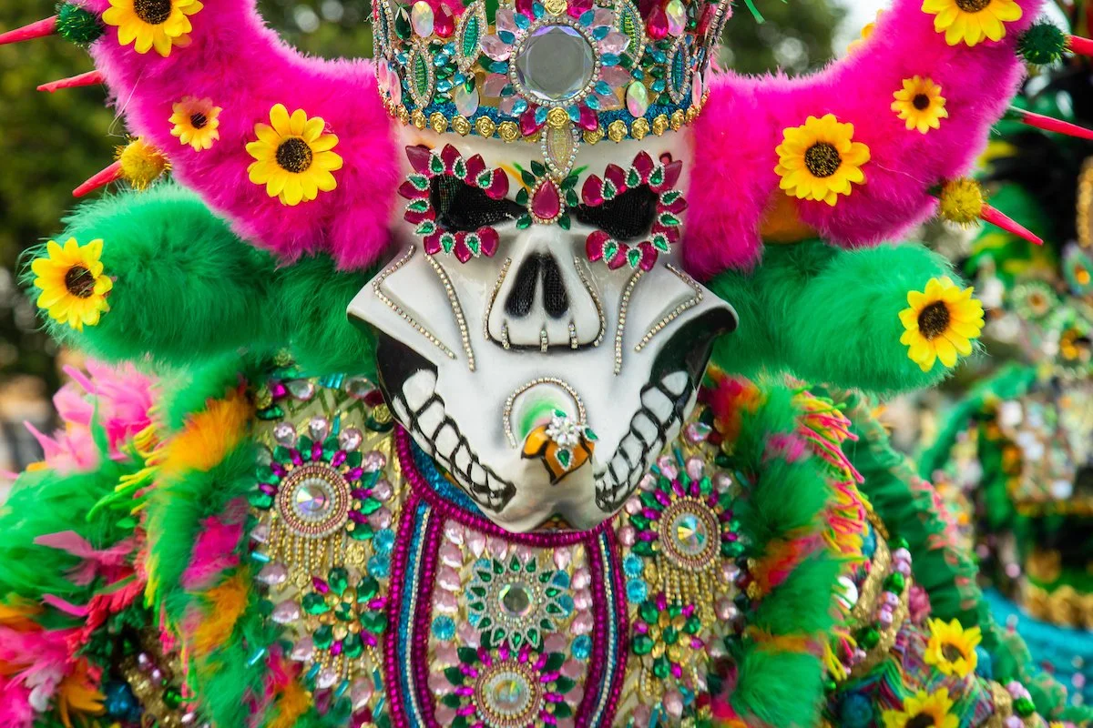 Colorful costume featuring a skull mask decorated with gemstones and flowers, with a headdress of pink and green fluffy feathers, and adornments with sunflowers and beads.