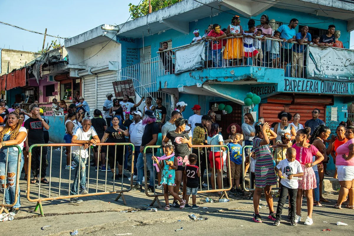 Crowd of people gathered on a street outside a blue building, with some standing behind barriers and others on the balcony, under a banner.