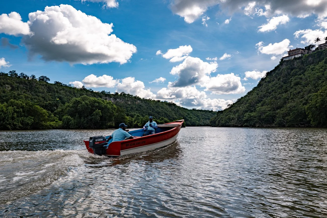 A boat with three men sailing on a river surrounded by green hills and a partly cloudy sky.