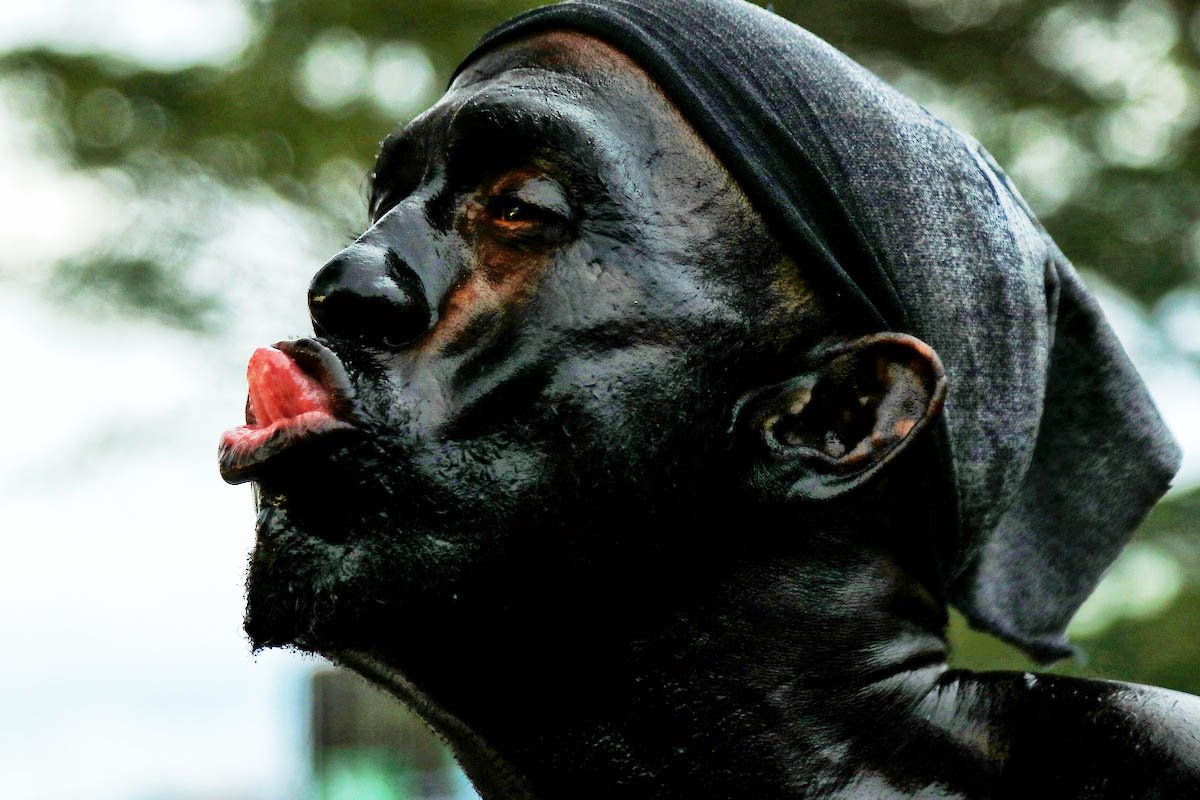 A person with a painted face making a silly expression, with their tongue out, wearing a dark shirt and a gray head covering, against a blurred outdoor background.