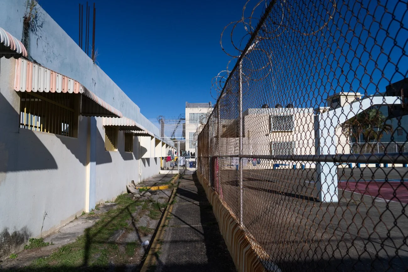 A narrow outdoor corridor between a white building with barred windows and a chain-link fence topped with barbed wire, with a sports court visible on the right and a clear blue sky overhead.