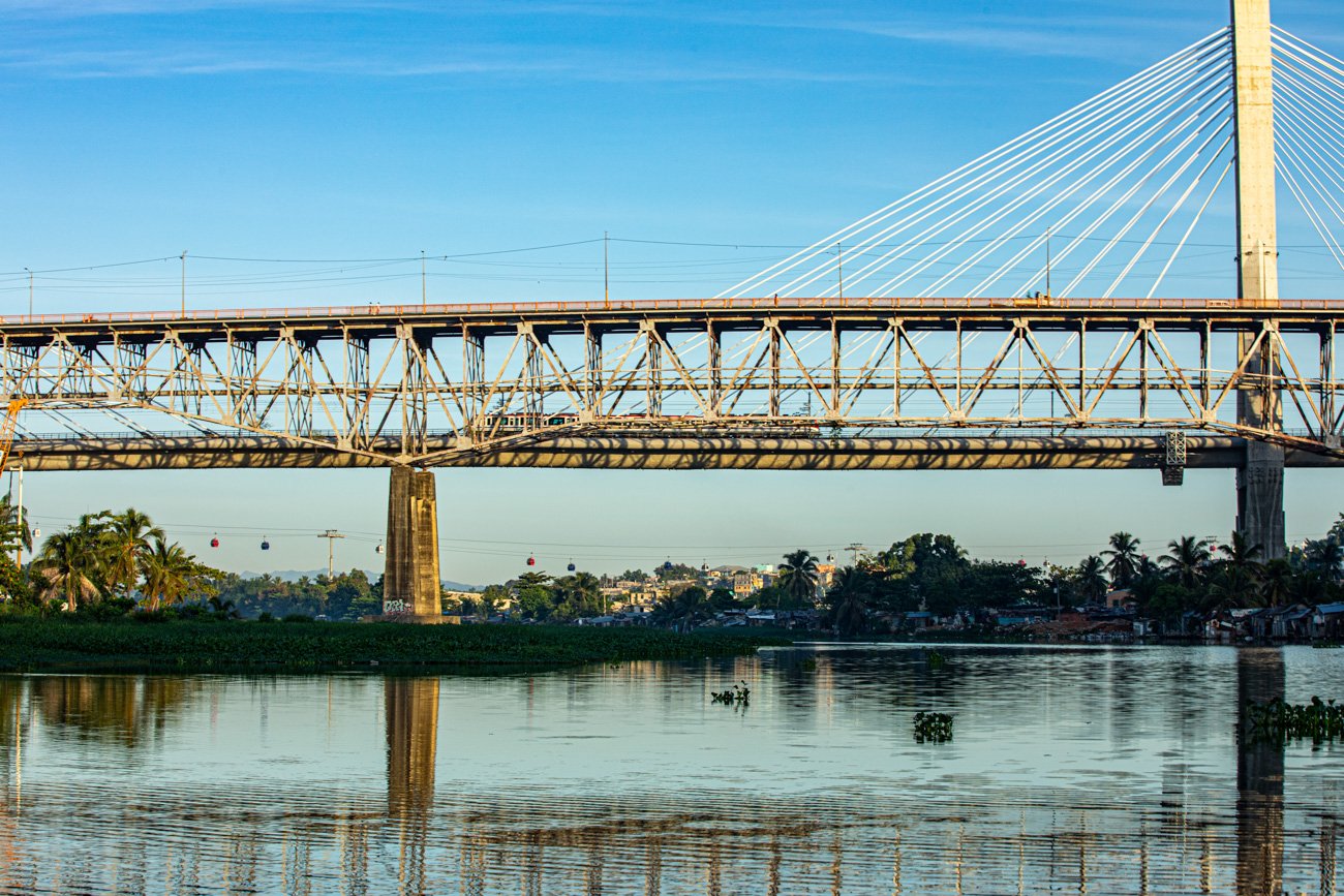 A large bridge crossing a body of water with a cityscape and cable cars in the background under a blue sky.
