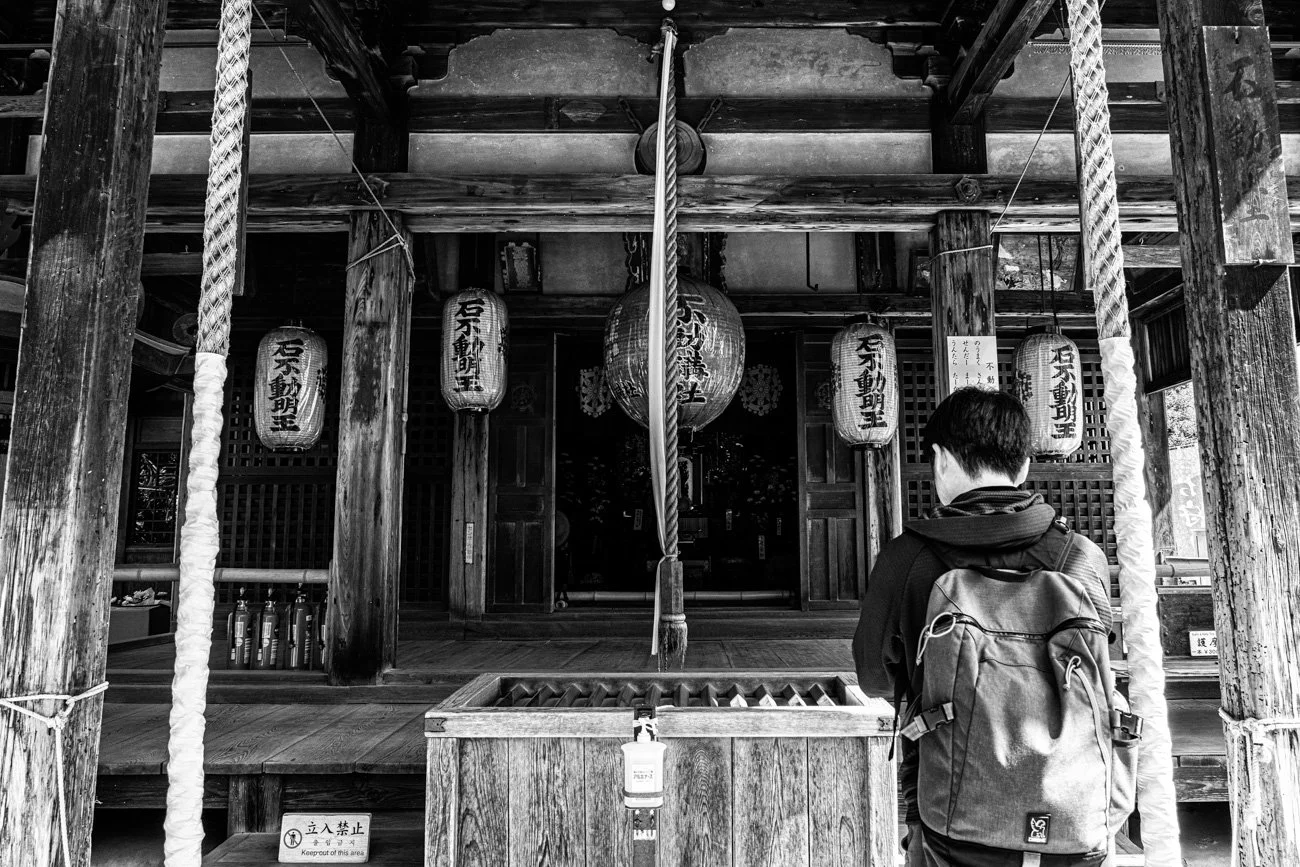 A person with a backpack standing in front of a traditional Japanese shrine, which features wooden beams, lanterns, and a hanging rope for prayer.