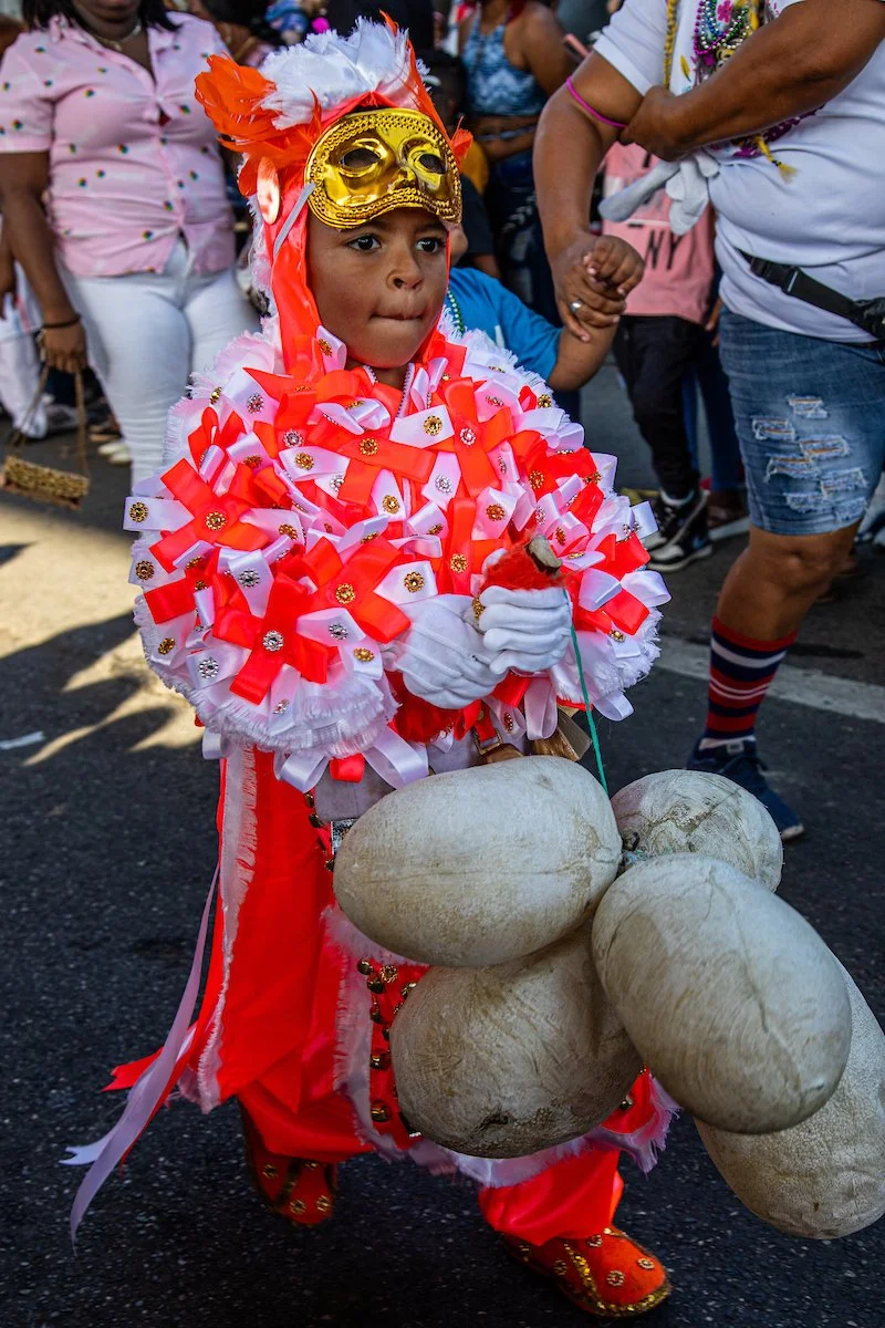 A young boy dressed in colorful costume wearing a golden mask with feathers, holding large stone-shaped objects, at a festive parade with other spectators in the background.