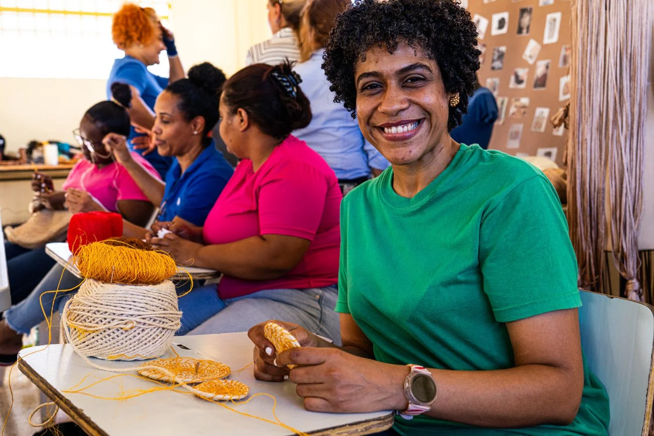 Smiling woman in green shirt sitting at table with yarn and knitting project, surrounded by women in colorful shirts in a craft or social setting.