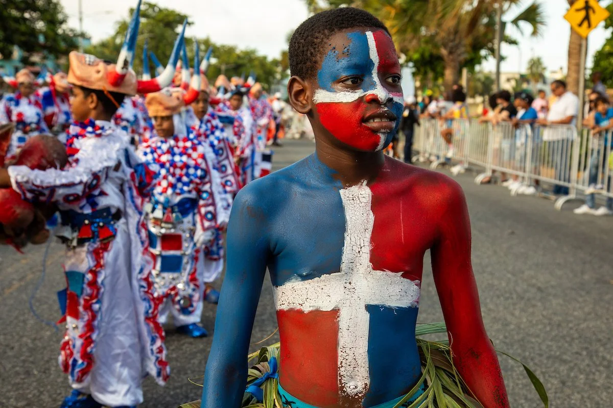 A young boy with face paint and body paint resembling the flags of France and Norway at a parade or celebration, with other costumed participants and spectators in the background.