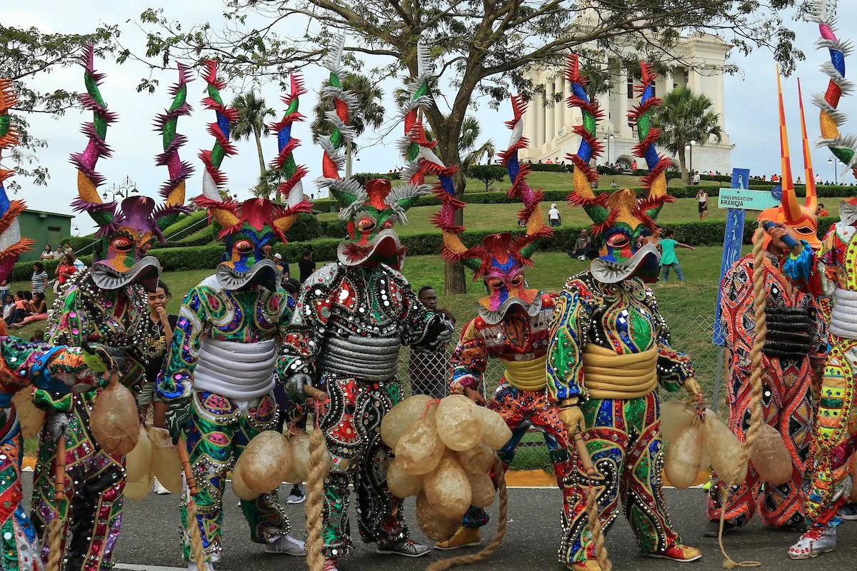 People dressed in colorful traditional costumes with masks and large headdresses, participating in a cultural parade or festival.