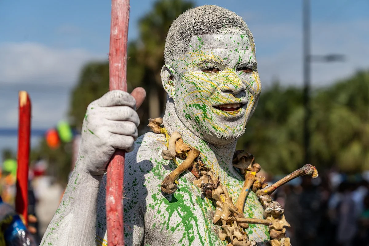 Person with painted face holding a red staff, wearing a necklace made of mushrooms and bones, attending an outdoor event with other people and trees in the background.
