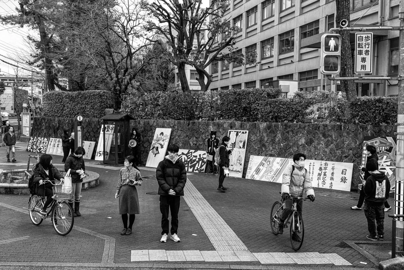 People waiting to cross the street at a pedestrian crosswalk in an urban area, with bicycles, posters, and Japanese signs on the surroundings.