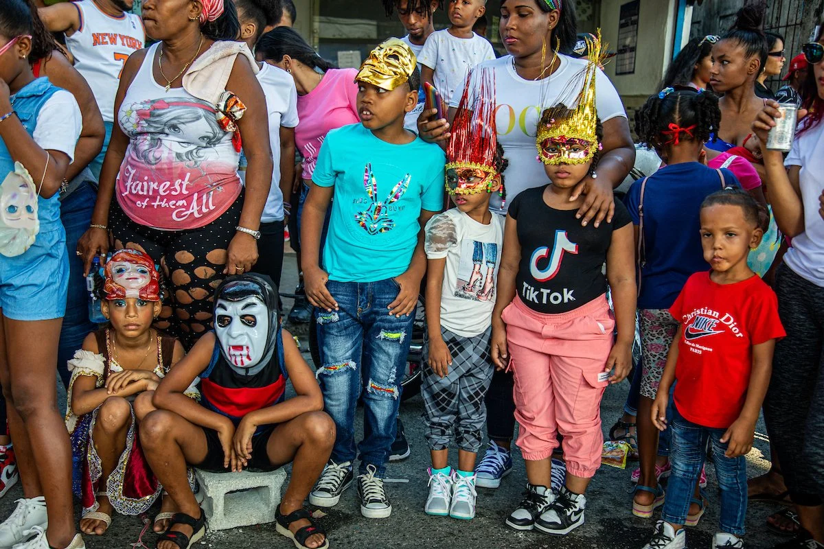 A group of children and adults gathered outdoors during a festival or parade, with some children wearing masks and costume accessories, and others in casual clothing.