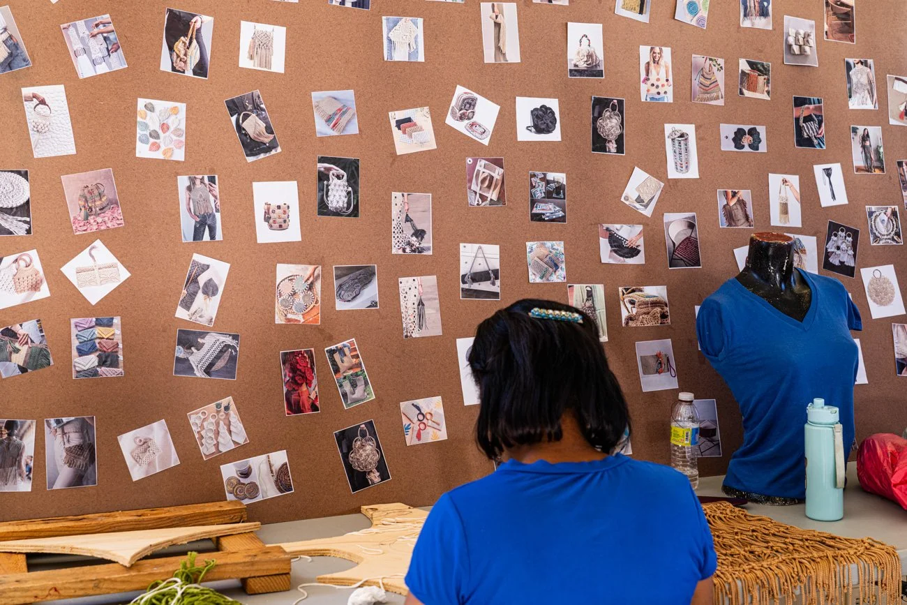 A woman working at a craft or design table with a corkboard wall behind displaying many photos of jewelry, clothing, and fabric patterns. She wears a blue shirt and has short dark hair. There is a mannequin with a blue shirt, water bottle, and red ba