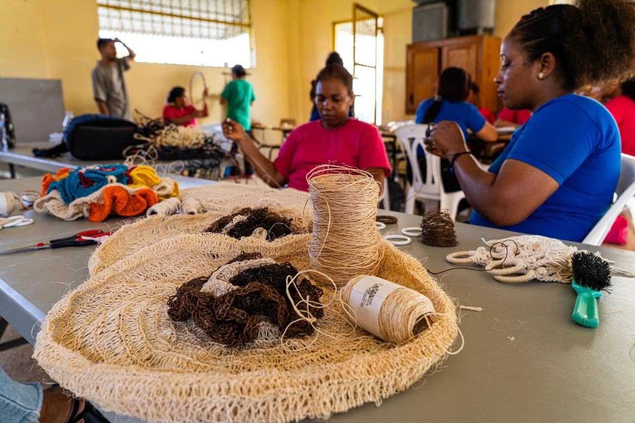 People working on crochet and textile crafts in a community room with yarn, scissors, and tools on a table.
