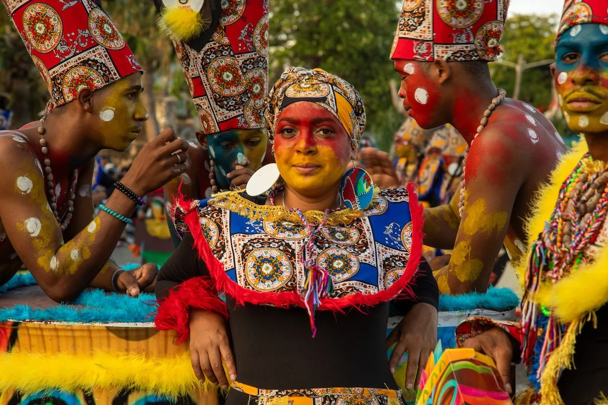 A group of people dressed in colorful traditional attire with face paint, jewelry, and headpieces, participating in a cultural festival or celebration outdoors.