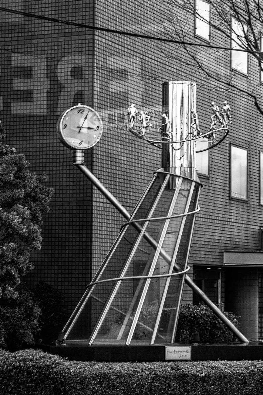 A large outdoor sculpture of a vintage street clock with a glass enclosure, supported by a metal pole. The clock shows 2:15. Attached to the pole is a miniature roller coaster with small cars, set against a brick building background.