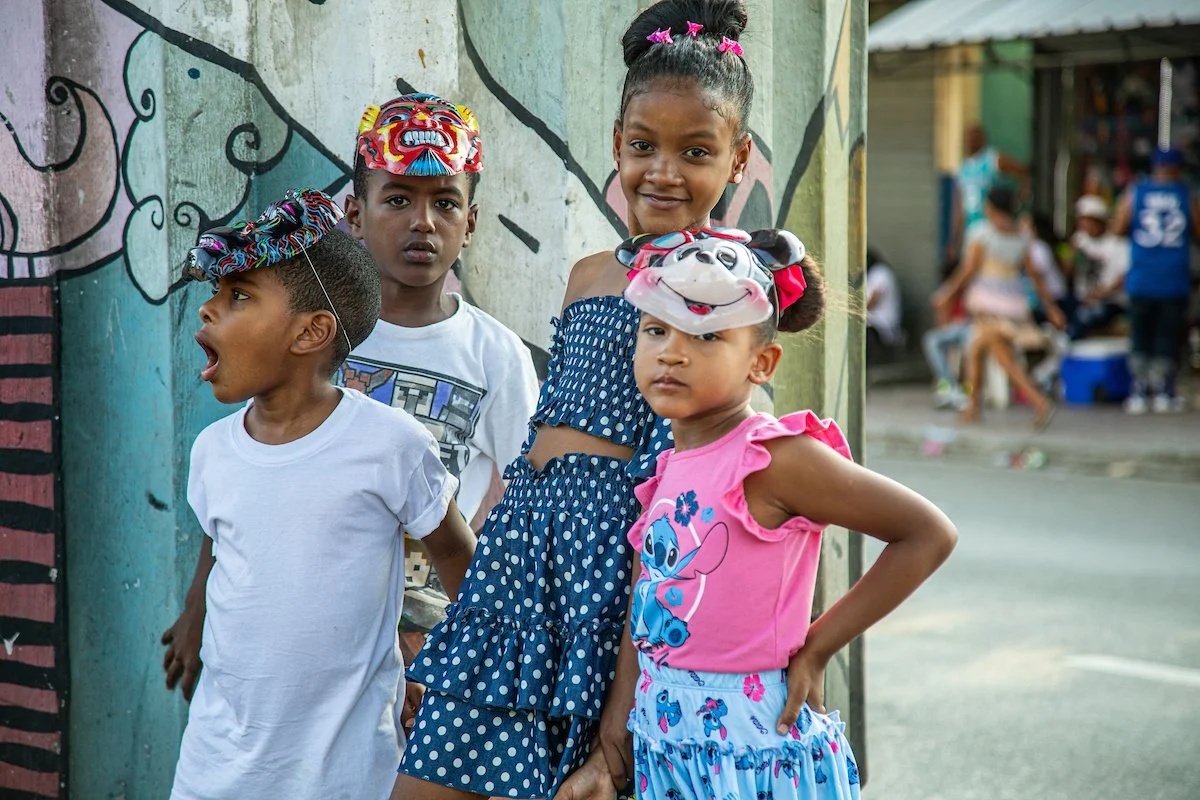 Four children standing outdoors in front of a colorful mural, wearing party masks, with a lively street scene in the background.
