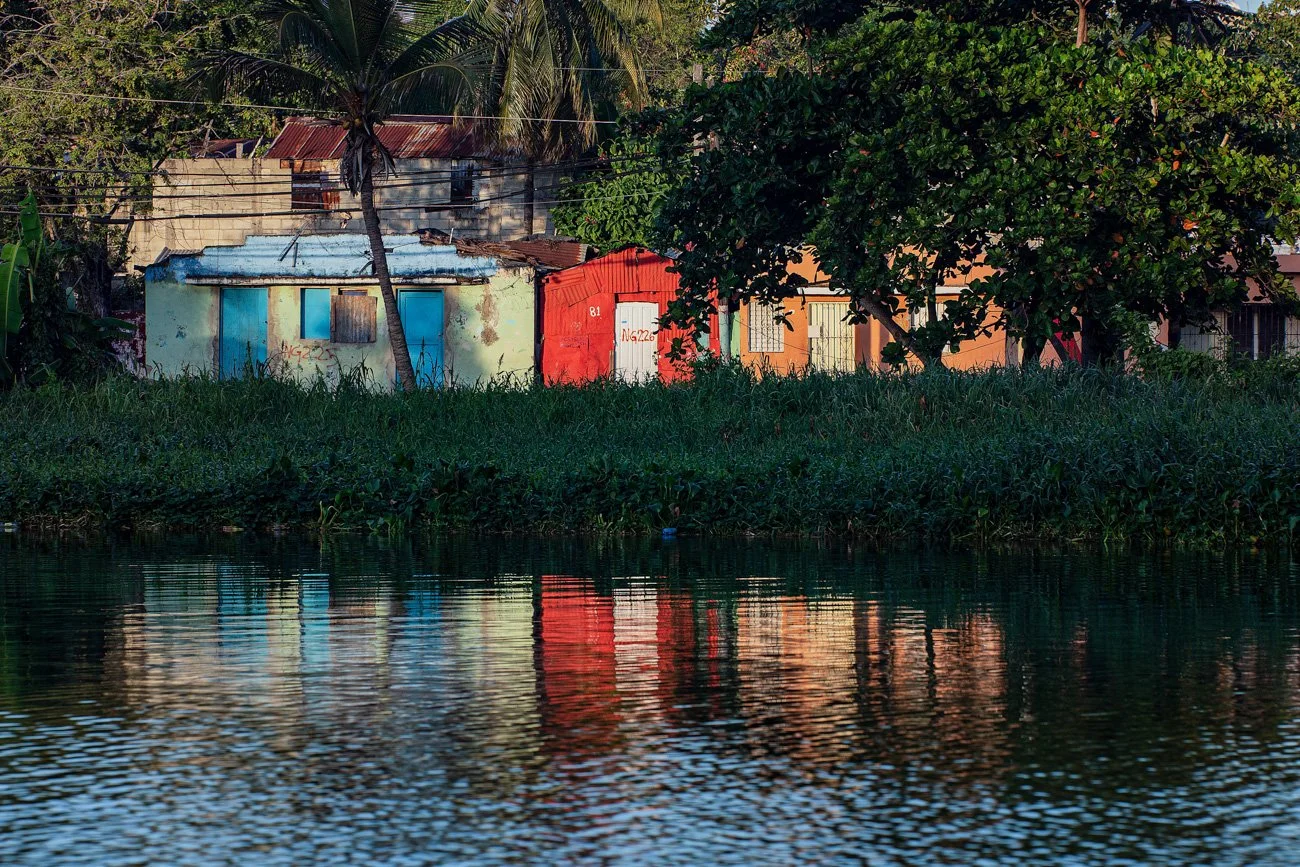 Colorful houses with tropical trees along a riverbank, with their reflections visible in the water.